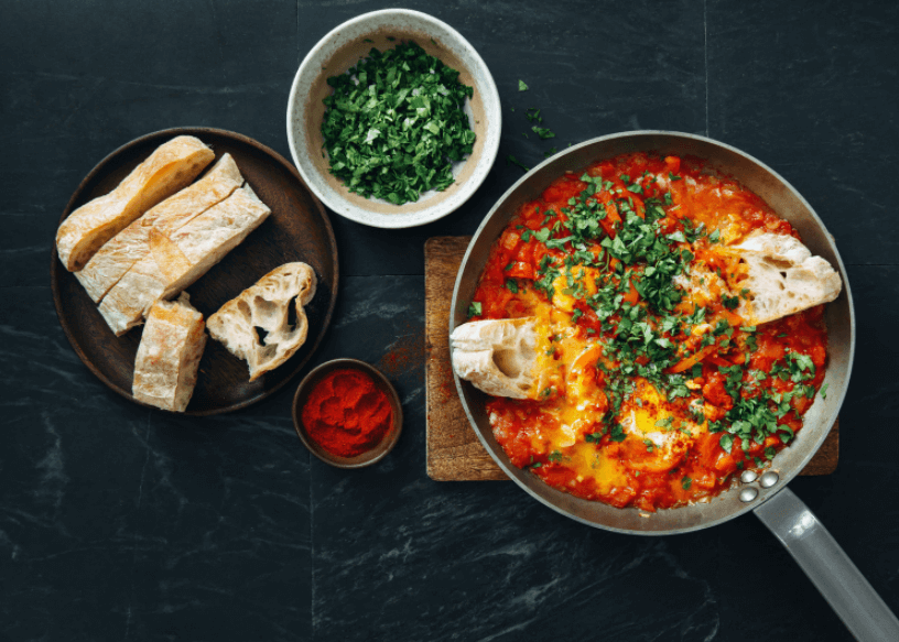 A pan with shakshouka poached fish served with bread and topped with fresh, green herbs.