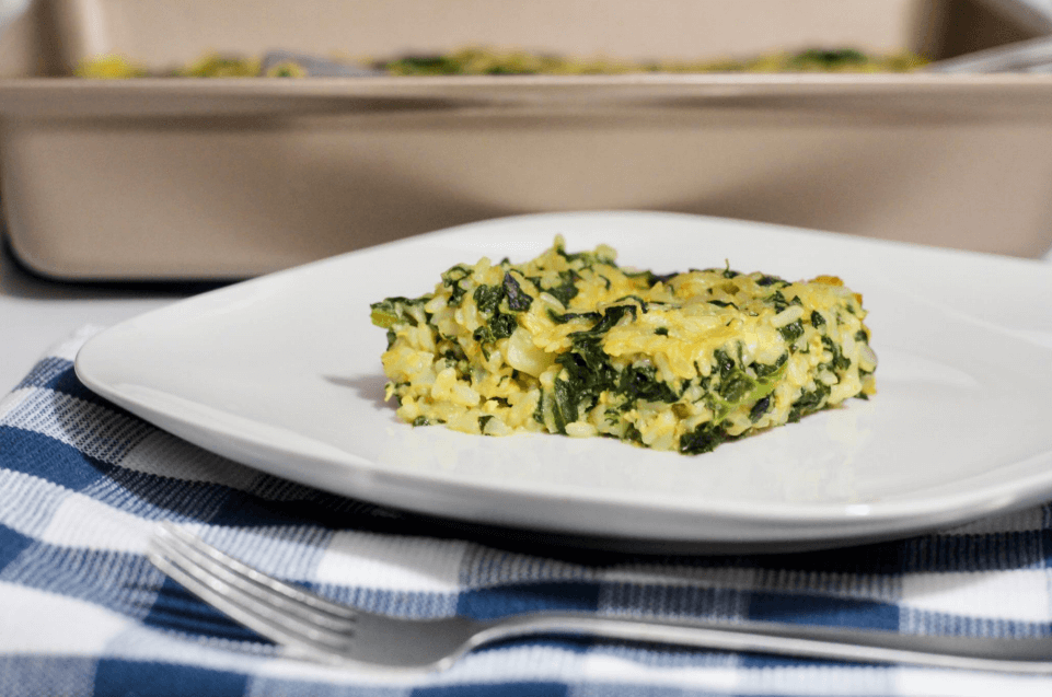A white plate with a single slice of spinach-rice casserole on top of a blue and white checkered cloth.