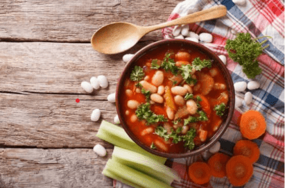 Tuscan vegetable soup in a dark bowl on top of a red and blue plaid cloth surrounded by sliced carrots, celery, and a wooden spoon.