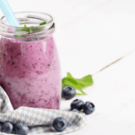 Creamy blueberry shake in a clear, glass jar next to a grey cloth with a white background.