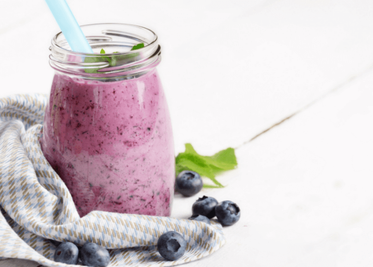 Creamy blueberry shake in a clear, glass jar next to a grey cloth with a white background.