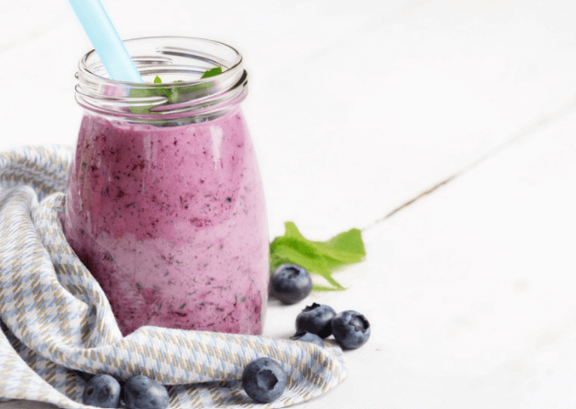 Creamy blueberry shake in a clear, glass jar next to a grey cloth with a white background.