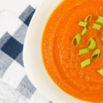 Bright, orange bowl of curried carrot soup topped with scallions on a blue and white checkered cloth.