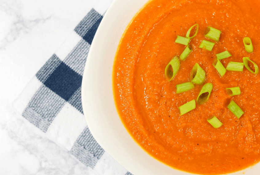 Bright, orange bowl of curried carrot soup topped with scallions on a blue and white checkered cloth.