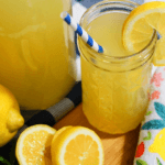 A tall glass jar filled with honey-sweetened lemonade garnished with a lemon slice. There is a blue- and white-striped straw in the glass and the glass is surrounded by lemons.