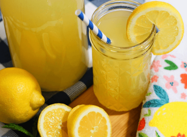 A tall glass jar filled with honey-sweetened lemonade garnished with a lemon slice. There is a blue- and white-striped straw in the glass and the glass is surrounded by lemons.