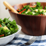 A small white bowl filled with massaged kale salad on top of a blue and white checkered cloth. A large wooden bowl of kale salad is in the background.