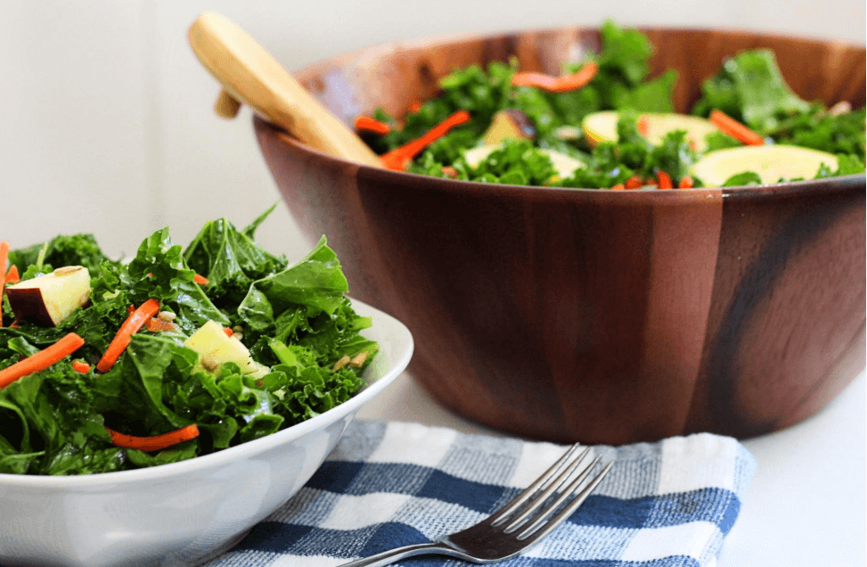 A small white bowl filled with massaged kale salad on top of a blue and white checkered cloth. A large wooden bowl of kale salad is in the background.