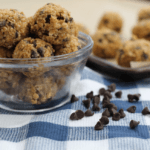 A small glass dish filled with no-bake chocolate chip bites. Chocolate chips are scattered on the blue- and white-checkered cloth next to the dish.