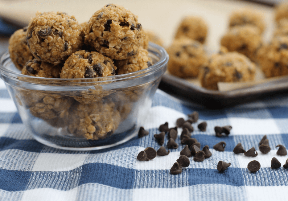 A small glass dish filled with no-bake chocolate chip bites. Chocolate chips are scattered on the blue- and white-checkered cloth next to the dish.