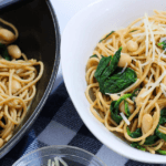 A white bowl with pasta, garlic, spinach, and white beans next to a black serving pan.