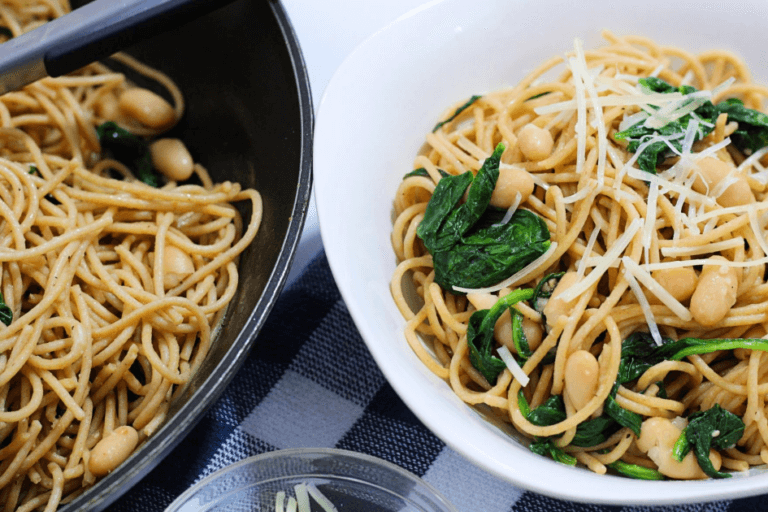 A white bowl with pasta, garlic, spinach, and white beans next to a black serving pan.