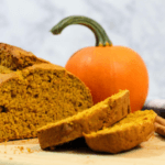 A loaf of pumpkin bread next to a small, orange pumpkin on a wooden cutting board.