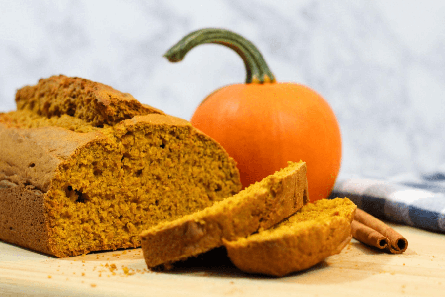 A loaf of pumpkin bread next to a small, orange pumpkin on a wooden cutting board.