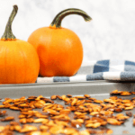 A baking pan full of roasted pumpkin seeds with two small, orange pumpkins in the background.