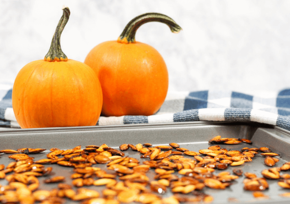 A baking pan full of roasted pumpkin seeds with two small, orange pumpkins in the background.