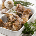 White baking dish with roasted rabbit. Surrounded by herbs and garlic.