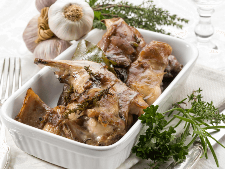 White baking dish with roasted rabbit. Surrounded by herbs and garlic.