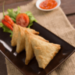 Black rectangular plate with four triangular sambusa and lettuce and tomato garnish on the side.