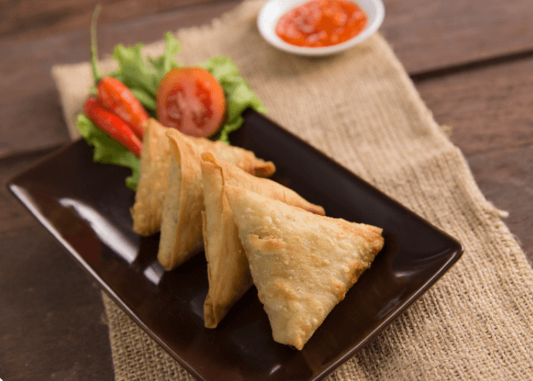 Black rectangular plate with four triangular sambusa and lettuce and tomato garnish on the side.