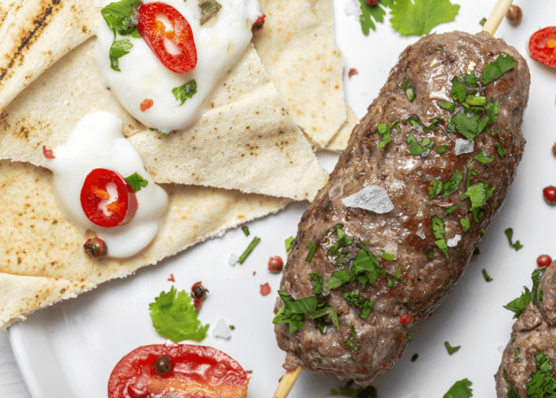 One kefta next to pieces of flatbread, white sauce, and slices of tomato.