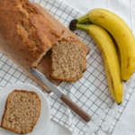 One loaf of banana bread with two cut pieces next to two bananas.