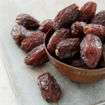 A bowl full of medjool dates with some dates scattered on the left side.