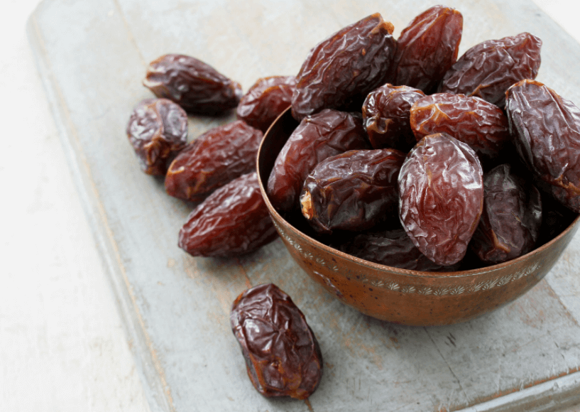 A bowl full of medjool dates with some dates scattered on the left side.