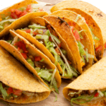 Several beef tacos in hard taco shells on a round wooden platter filled with lettuce and tomatoes.