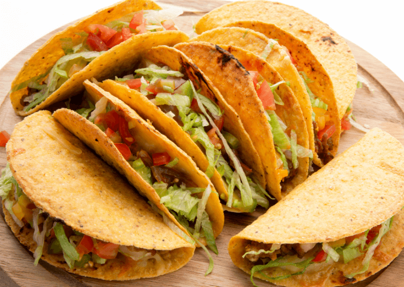 Several beef tacos in hard taco shells on a round wooden platter filled with lettuce and tomatoes.