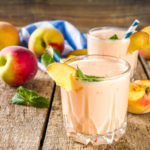 Two peach smoothies in a clear glass surrounded by whole peaches on a wooden surface.