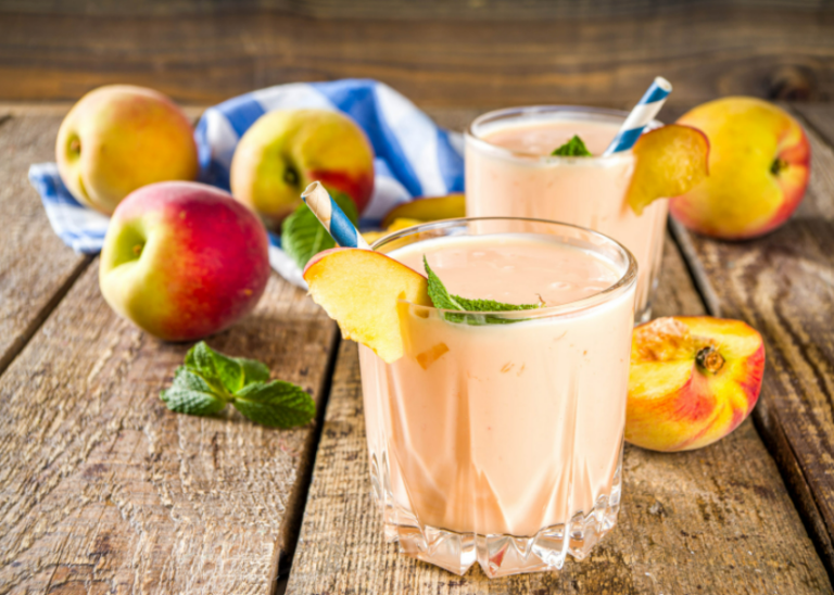 Two peach smoothies in a clear glass surrounded by whole peaches on a wooden surface.