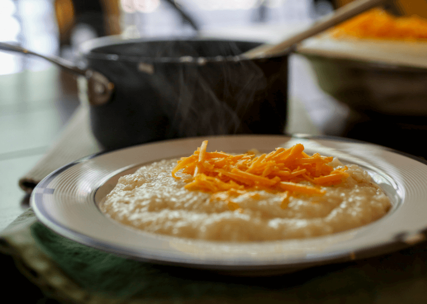 A shallow bowl of savory farina topped with bright orange shredded cheese and a saucepan in the background.