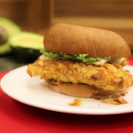 Southern-battered catfish sandwich on a white plate on top of a red surface. Sliced avocado in the background.