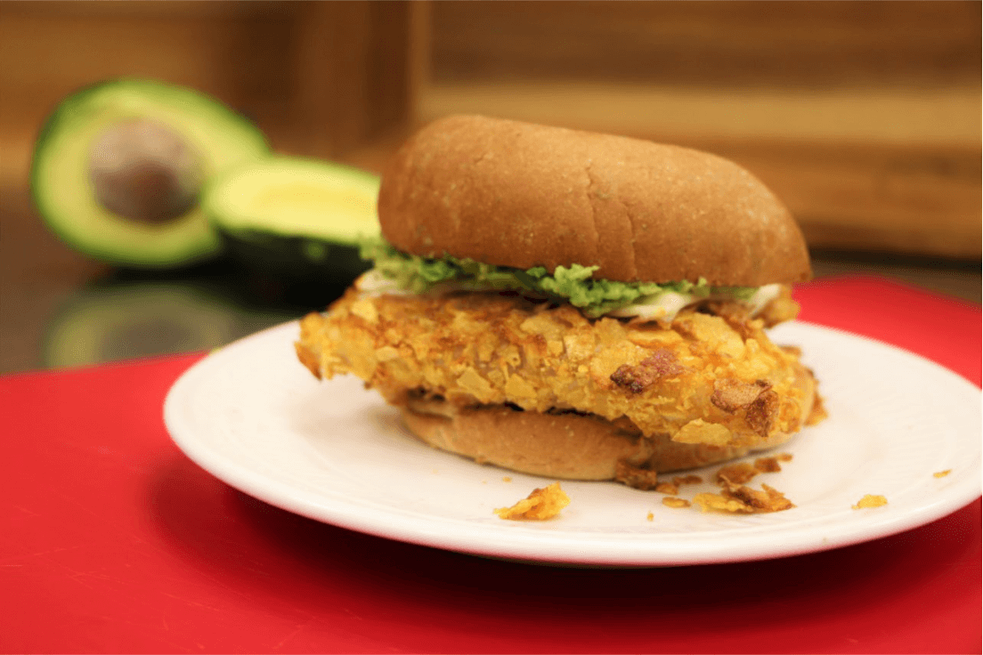 Southern-battered catfish sandwich on a white plate on top of a red surface. Sliced avocado in the background.