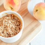 A small white dish with peach crisp on top of a wooden platter with two whole peaches next to it.