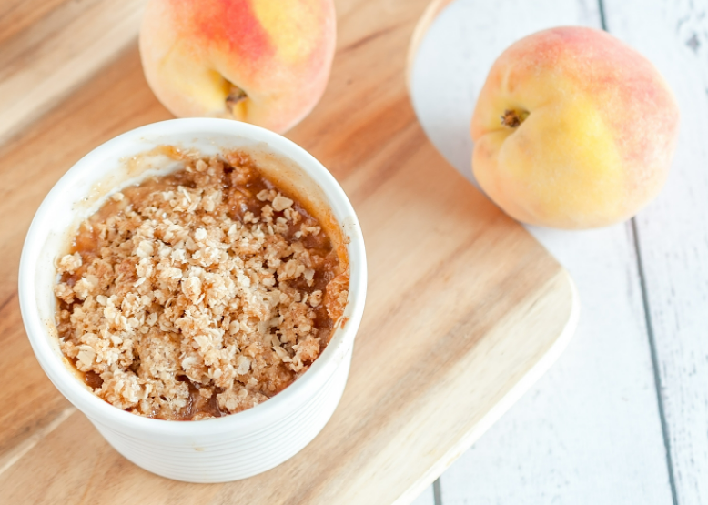 A small white dish with peach crisp on top of a wooden platter with two whole peaches next to it.