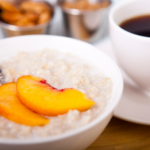 Peaches and cream oatmeal topped with two peach slices in a white bowl with a cup of coffee in the background.