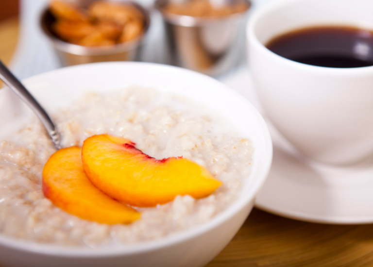Peaches and cream oatmeal topped with two peach slices in a white bowl with a cup of coffee in the background.