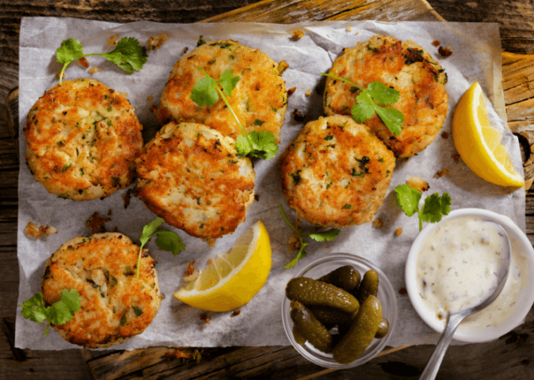 Hake fish cakes on served with lemon wedges, pickles, and a small cup of tartar sauce.