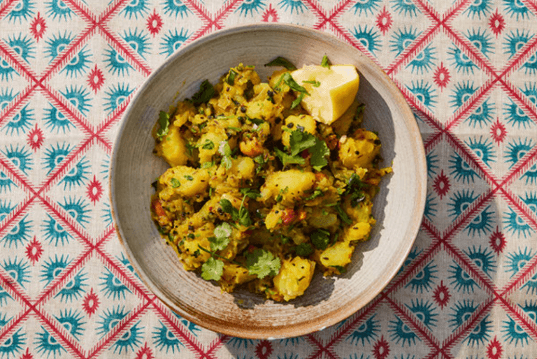 A bowl of aloo masala with a colorful, patterned background. Garnished with fresh green herbs and a slice of lemon.