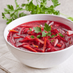 A white bowl full of borscht with some green herbs in the background.