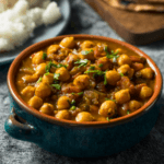 A small blue bowl filled with chana masala, with rice and other side dishes in the background.