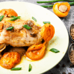 A white plate with a chicken thigh and apricot halves topped with green herbs.