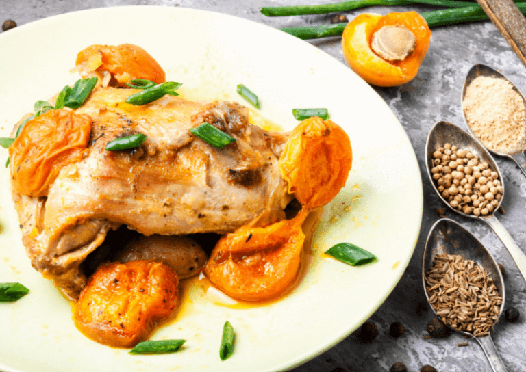 A white plate with a chicken thigh and apricot halves topped with green herbs.