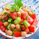 A bright bowl of chickpea salad with tomatoes, cucumbers, and fresh herbs on top of a blue, patterned cloth.