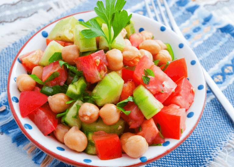 A bright bowl of chickpea salad with tomatoes, cucumbers, and fresh herbs on top of a blue, patterned cloth.