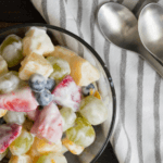 Fruit with yogurt topping in a glass bowl next to a striped cloth with two spoons.