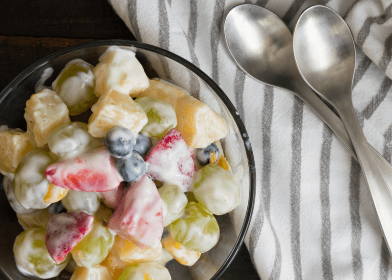 Fruit with yogurt topping in a glass bowl next to a striped cloth with two spoons.