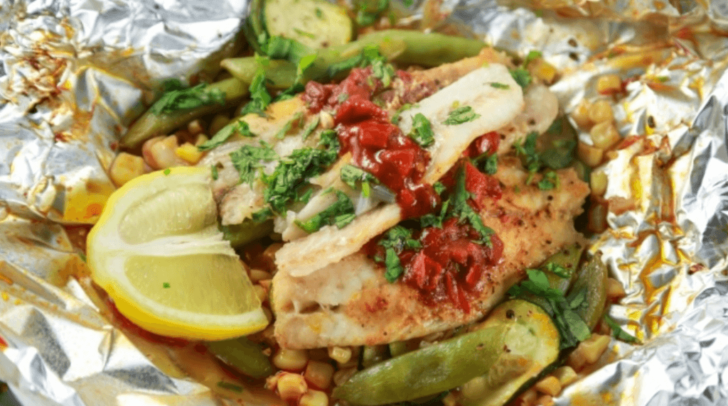 Grilled walleye in aluminum foil with vegetables and a lemon wedge. Topped with harissa sauce.